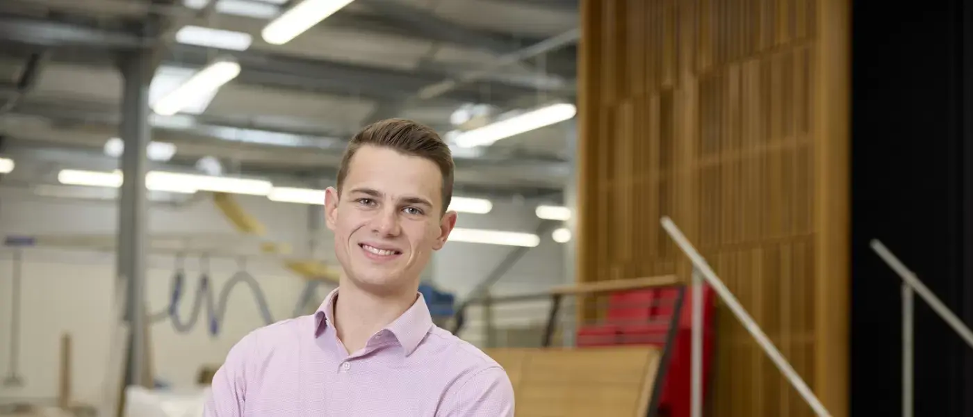 A young man wearing a light pink shirt stands confidently in a joinery workshop smiling with arms crossed The background includes woodworking tools timber materials and modern workshop equipment
